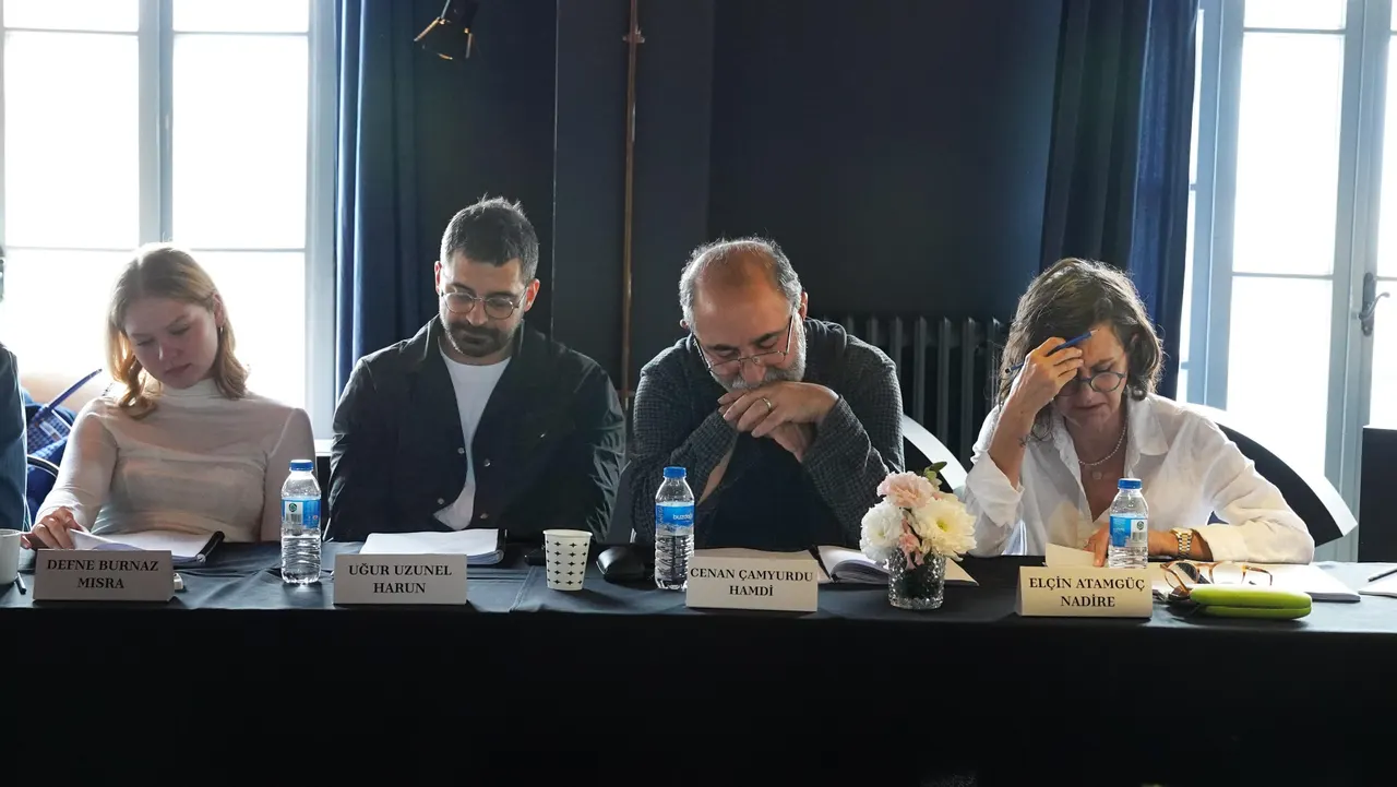 Oyuncular isim ve rol kartlarının bulunduğu okuma provası masasında oturuyor.